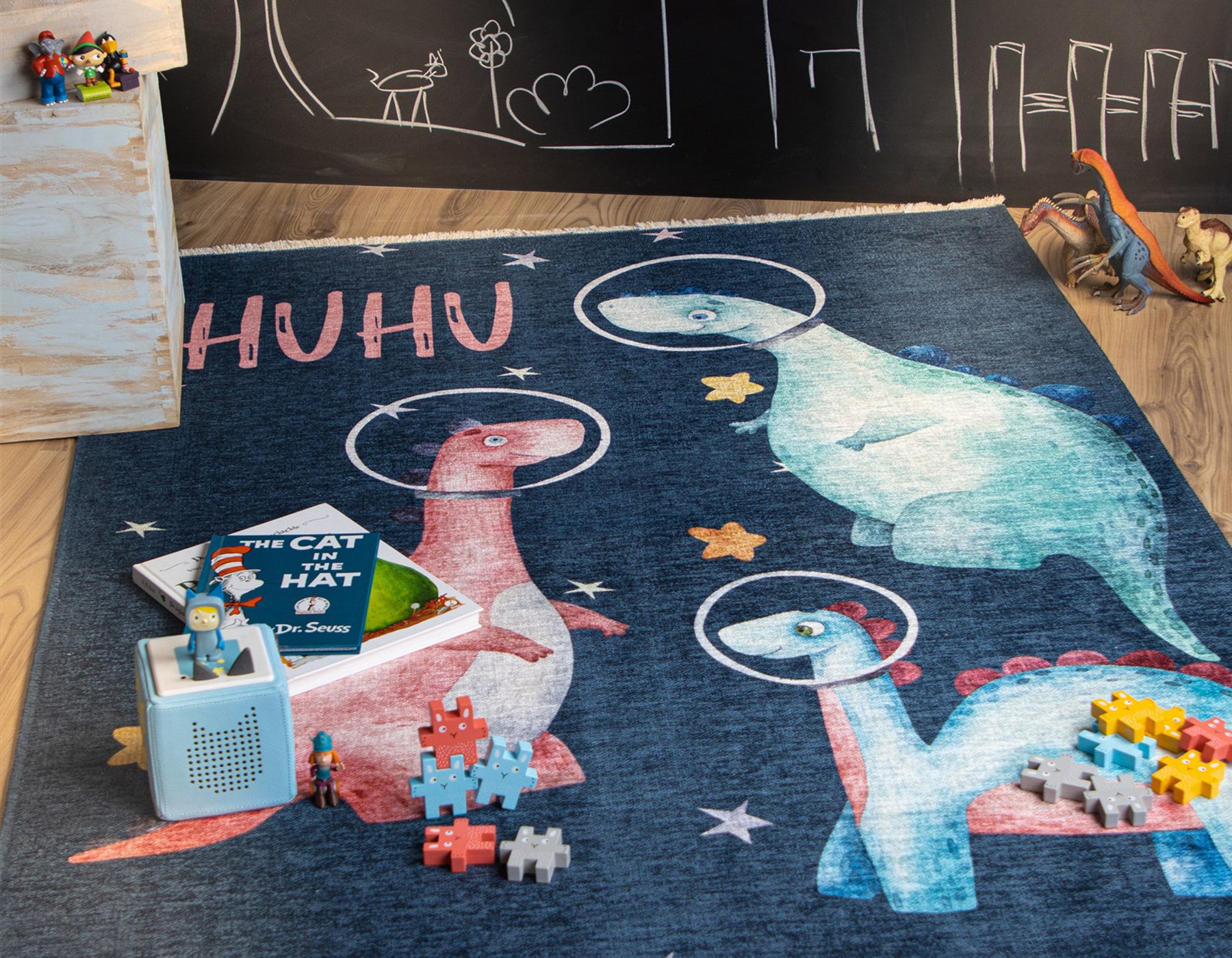 Kurzflor Kinderteppich Joanna 627 Huhu aus Polyester in blau mit bunten kindlichem Dinos die eine Art Raumhelm tragen mit Sternen und einem Schriftzug Huhu.